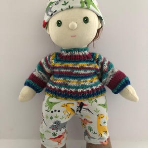 May include: A handmade doll wearing a colorful striped sweater, dinosaur-print pants, and a matching hat. The doll has brown hair, green eyes, and brown shoes. The doll is approximately 12 inches tall.