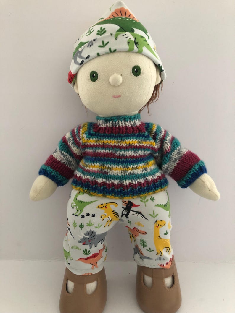 May include: A handmade doll wearing a colorful striped sweater, dinosaur-print pants, and a matching hat. The doll has brown hair, green eyes, and brown shoes. The doll is approximately 12 inches tall.