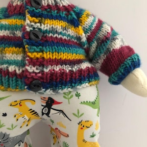 May include: A doll wearing a colorful, striped knit cardigan with three black buttons. The doll also wears white pants patterned with various cartoon dinosaurs in yellow, green, and gray.