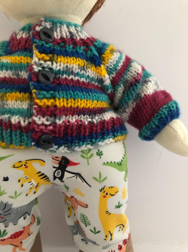May include: A doll wearing a colorful, striped knit cardigan with three black buttons. The doll also wears white pants patterned with various cartoon dinosaurs in yellow, green, and gray.