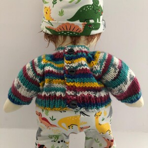 May include: A doll wearing a colorful knit sweater with black buttons, white pants with dinosaur patterns, and a white hat with dinosaur illustrations. The sweater has stripes of teal, purple, yellow, and white.