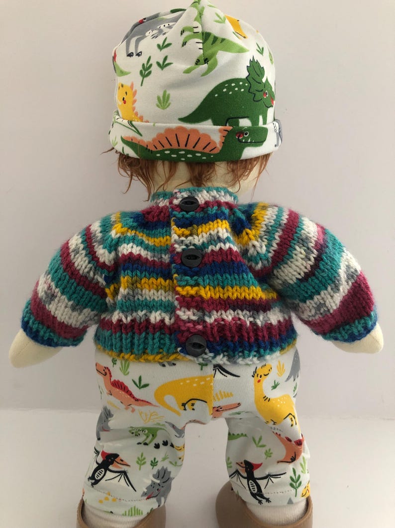 May include: A doll wearing a colorful knit sweater with black buttons, white pants with dinosaur patterns, and a white hat with dinosaur illustrations. The sweater has stripes of teal, purple, yellow, and white.