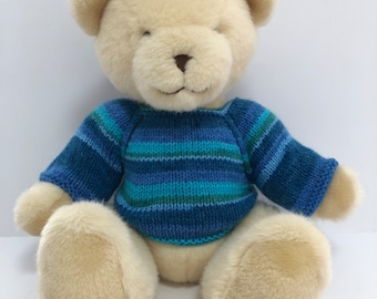 Può includere: Un orsacchiotto color crema che indossa un maglione a righe blu e verdi. L'orso è seduto con le zampe davanti a sé.