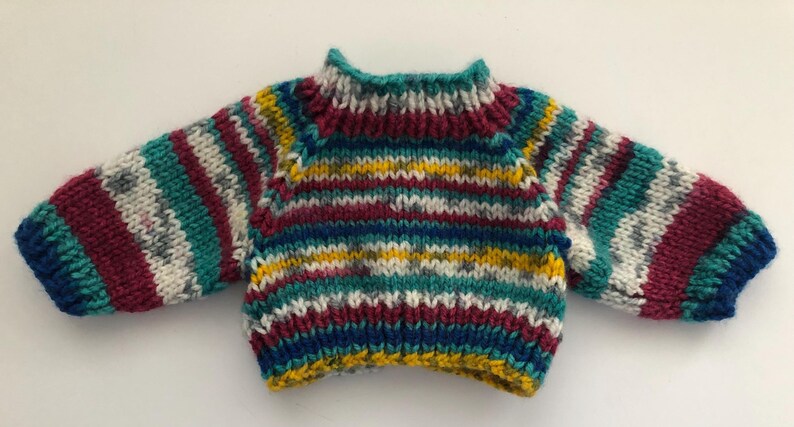 May include: A small, hand-knitted sweater in a variety of colors, including teal, burgundy, yellow, and white. The sweater has a high neck and long sleeves. The sleeves and bottom hem are a darker blue. The sweater is approximately 10 inches wide.