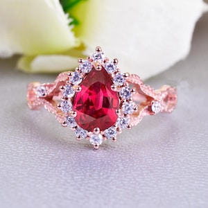May include: Rose gold ring featuring a pear-shaped, vibrant red gemstone surrounded by small white stones. The ring's band has an intricate, openwork design. The ring is set against a soft, light gray background with white floral accents.