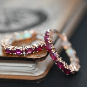 Op de afbeelding: Twee roségouden ringen met ovaal geslepen robijn edelstenen. Eén ring is scherp, met de heldere rode kleur van de robijnen. De ringen liggen op een houten oppervlak met een vage achtergrond.