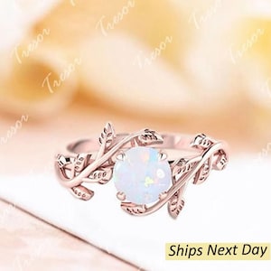 May include: Rose gold ring featuring a round, white opal gemstone. The ring's band is adorned with delicate leaf and vine detailing, creating an elegant, nature-inspired design. The ring is a beautiful piece of jewelry.