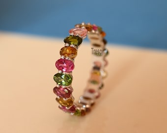 May include: A silver ring with a band of oval-shaped gemstones in shades of pink, green, and yellow. The ring is set on a white background.