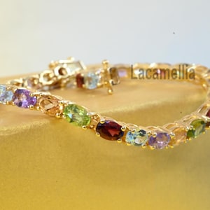 May include: A gold-toned bracelet featuring a series of oval-cut multi-colored gemstones including red, purple, green, and light blue stones set in a metal chain. The word Lacamellia is printed in black text near the center of the bracelet.