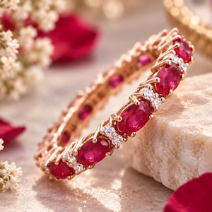 May include: A rose gold eternity band featuring a repeating pattern of oval-cut vibrant red rubies alternating with pairs of small round-cut clear white diamonds set in a continuous circular metal frame.