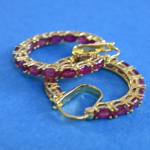 Könnte beinhalten: Goldfarbene Creolen-Ohrringe mit einer durchgehenden Reihe oval geschliffener, tiefroter Edelsteine. Die Ohrringe sind vor einem leuchtend blauen Hintergrund platziert, der das Design und die Farbe hervorhebt. Ein klassisches Schmuckstück.