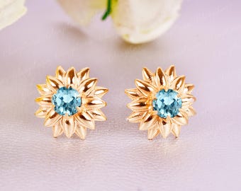 May include: A pair of gold-colored stud earrings. Each earring features a sunflower design with a light blue gemstone center. The petals of the sunflowers are detailed and pointed. The earrings are set against a soft, light background.