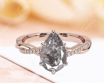 Puede incluir: Un anillo de compromiso de diamante gris en forma de pera engastado en oro rosa con una banda retorcida adornada con pequeños diamantes.