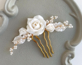 May include: A white floral hair comb featuring a large rose, smaller buds, and pearl accents. The gold-toned metal comb has three curved prongs. The design includes white leaves and small pearl-like beads, perfect for bridal or special occasion hairstyles.