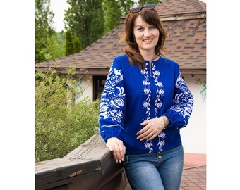 May include: A woman wearing a blue embroidered blouse with white floral designs and blue jeans. She is standing in front of a wooden structure with a brown roof.