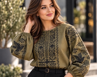 May include: An olive green long-sleeved blouse featuring intricate black geometric embroidery and bird motifs on the front and sleeves. The garment has a round neckline, gathered cuffs, and is paired with black trousers featuring a belt.