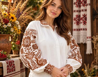 May include: A white long-sleeved blouse featuring intricate brown embroidery on the sleeves, cuffs, and collar. The design includes detailed floral and bird motifs, with a button-down front placket and a relaxed fit, showcasing traditional needlework.