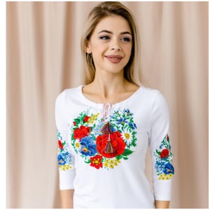 May include: White long-sleeved shirt with a floral embroidery design featuring red, blue, and yellow flowers. The embroidery is in the shape of a circle and has a tassel detail.