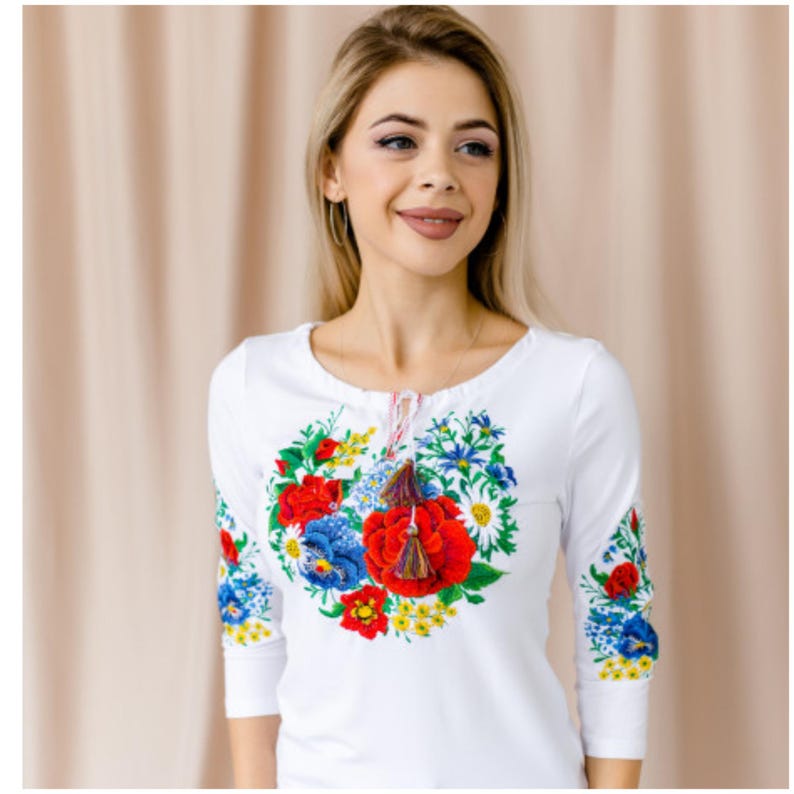 May include: White long-sleeved shirt with a floral embroidery design featuring red, blue, and yellow flowers. The embroidery is in the shape of a circle and has a tassel detail.
