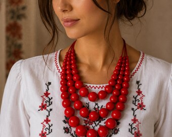 May include: A multi-strand necklace featuring four layers of glossy, round, bright red beads of varying sizes. The necklace is worn over a white long-sleeved shirt that displays intricate red and black floral embroidery along the front neckline.