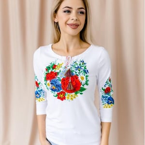 May include: White long-sleeved shirt with a floral embroidery design featuring red, blue, and yellow flowers. The shirt has a round neckline and a tassel detail.