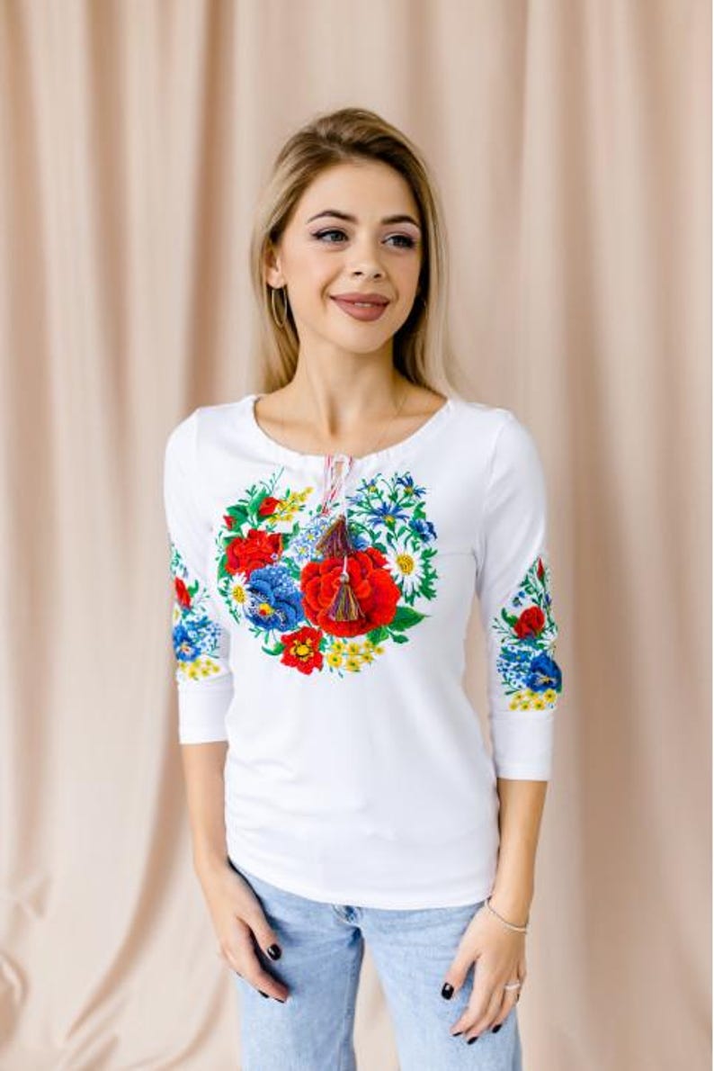 May include: White long-sleeved shirt with a floral embroidery design featuring red, blue, and yellow flowers. The shirt has a round neckline and a tassel detail.