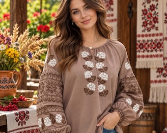 May include: A light brown long-sleeve blouse featuring intricate white and dark brown floral embroidery on the chest and sleeves. The garment has a round neckline, gathered cuffs, and a relaxed fit, paired with blue denim jeans.