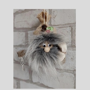 May include: A gray and white fuzzy gnome ornament with a burlap sack hat and a key hanging from its hand. The gnome is hanging on a white brick wall.