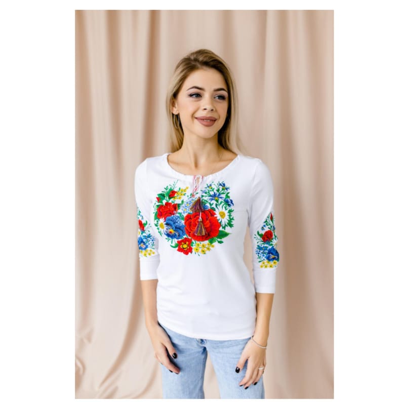 May include: White long-sleeved shirt with a floral embroidery design in red, blue, and yellow. The embroidery features a large bouquet of flowers with a red poppy in the center.