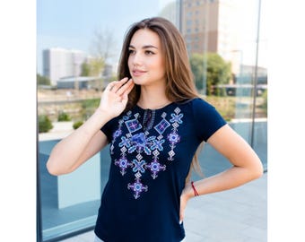 May include: Navy blue short-sleeve top with intricate floral embroidery in shades of blue and purple. The embroidered design is centered on the chest and neckline. The top has a round neckline with a small tie detail.