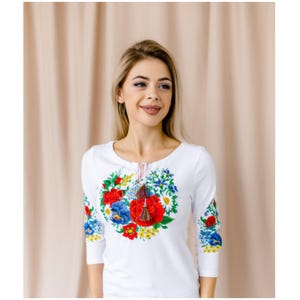 May include: White long-sleeved shirt with a floral embroidery design featuring red, blue, and yellow flowers. The embroidery is in the shape of a heart and has a tassel hanging from the center.