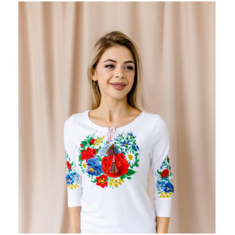 May include: White long-sleeved shirt with a floral embroidery design featuring red, blue, and yellow flowers. The embroidery is in the shape of a heart and has a tassel hanging from the center.