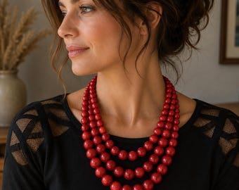May include: A multi-strand necklace featuring three layers of polished, round, deep red beads. The beads are uniform in size and arranged in a cascading design that rests against the collarbone, creating a bold and vibrant statement piece.
