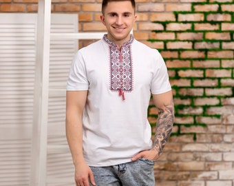 Peut inclure: Une chemise blanche à manches courtes avec un motif brodé traditionnel sur le devant. La chemise a un col montant et une coupe décontractée. La personne portant la chemise est debout devant un mur de briques avec de la mousse verte.
