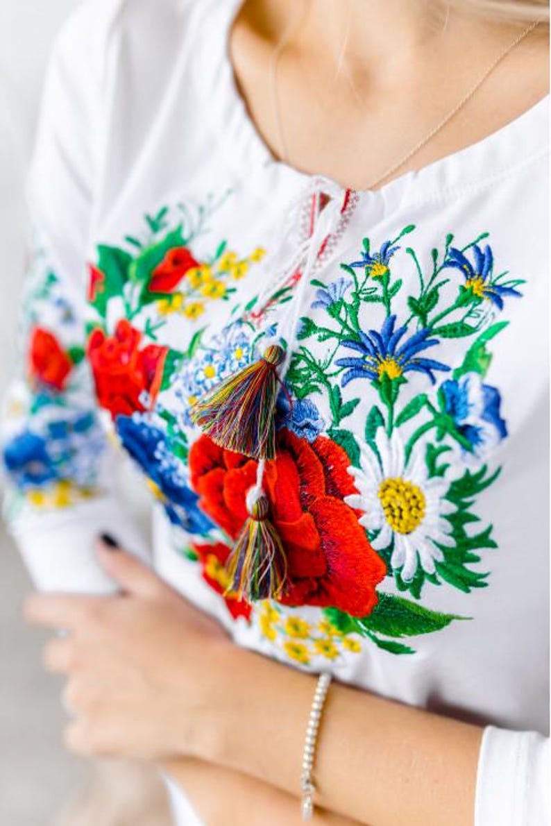 May include: White embroidered blouse with a floral design featuring red, blue, yellow, and green flowers. The blouse has a tie at the neck and tassels.