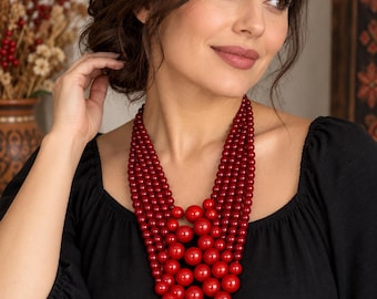 May include: A multi-strand necklace featuring glossy, deep red spherical beads of varying sizes. The strands are arranged in a cascading, layered design that sits against the chest, with larger beads concentrated toward the center of the necklace.