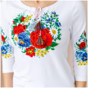 May include: White long-sleeved shirt with embroidered floral design in red, blue, yellow, and green. The embroidery features a large bouquet of flowers with tassels.