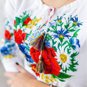 May include: White embroidered blouse with a floral design featuring red, blue, yellow, and green flowers. The blouse has a tie at the neck and tassels.
