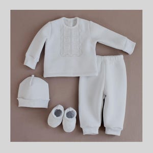 Op de afbeelding: Een witte baby outfit set met een shirt met lange mouwen, broek, muts en slofjes. Het shirt heeft een wit geborduurd ontwerp op de voorkant.