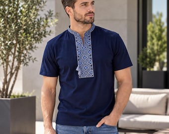 May include: A short-sleeved navy blue shirt featuring a stand-up collar and a central vertical panel with intricate blue and white geometric embroidery. The neckline includes a small slit and two hanging tassels at the base of the embroidery.