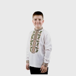 Può includere: Una camicia bianca a maniche lunghe con un motivo ricamato tradizionale che presenta foglie verdi e fiori marroni. La camicia ha un colletto alto e una chiusura a zip.