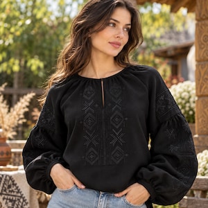 May include: A black long-sleeve blouse featuring a round neckline with a small vertical slit and intricate black geometric embroidery on the chest and sleeves. The garment has voluminous sleeves with gathered cuffs and a relaxed fit throughout.