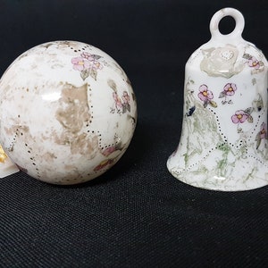 Op de afbeelding: Twee witte keramische ornamenten met een bloemenmotief. Een ornament is een bol met een gouden lint eraan bevestigd. Het andere ornament is een bel met een lus aan de bovenkant.