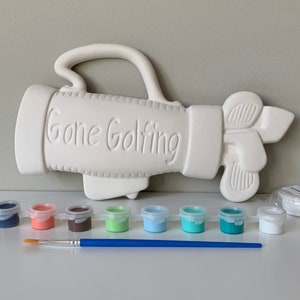 May include: A white ceramic golf bag with the words "Gone Golfing" on the side. The bag is accompanied by a set of seven small paint pots in various colors, a paintbrush, and a white ceramic disc.