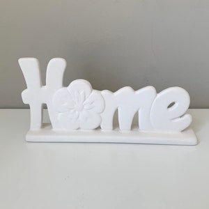 Puede incluir: Un letrero de cer&aacute;mica blanca para el hogar con un dise&ntilde;o floral. El letrero dice "Home".