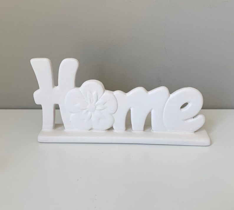 Puede incluir: Un letrero de cer&aacute;mica blanca para el hogar con un dise&ntilde;o floral. El letrero dice "Home".