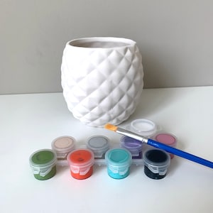 Puede incluir: Una maceta de cer&aacute;mica blanca con forma de pi&ntilde;a y superficie texturizada. La maceta est&aacute; rodeada de ocho peque&ntilde;os botes de pintura de varios colores, entre ellos verde, naranja, turquesa, negro, morado, gris, beige y blanco. Un pincel est&aacute; encima de los botes de pintura.