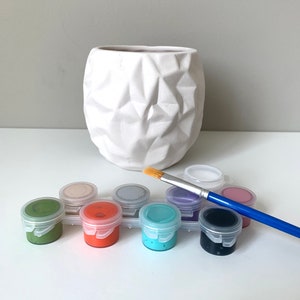 Puede incluir: Un macetero de cer&aacute;mica blanca con un dise&ntilde;o geom&eacute;trico, un juego de botes de pintura de varios colores y un pincel.