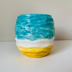 Puede incluir: Una taza de cer&aacute;mica con un esmalte azul, blanco y amarillo. La taza tiene una superficie ondulada y texturizada.