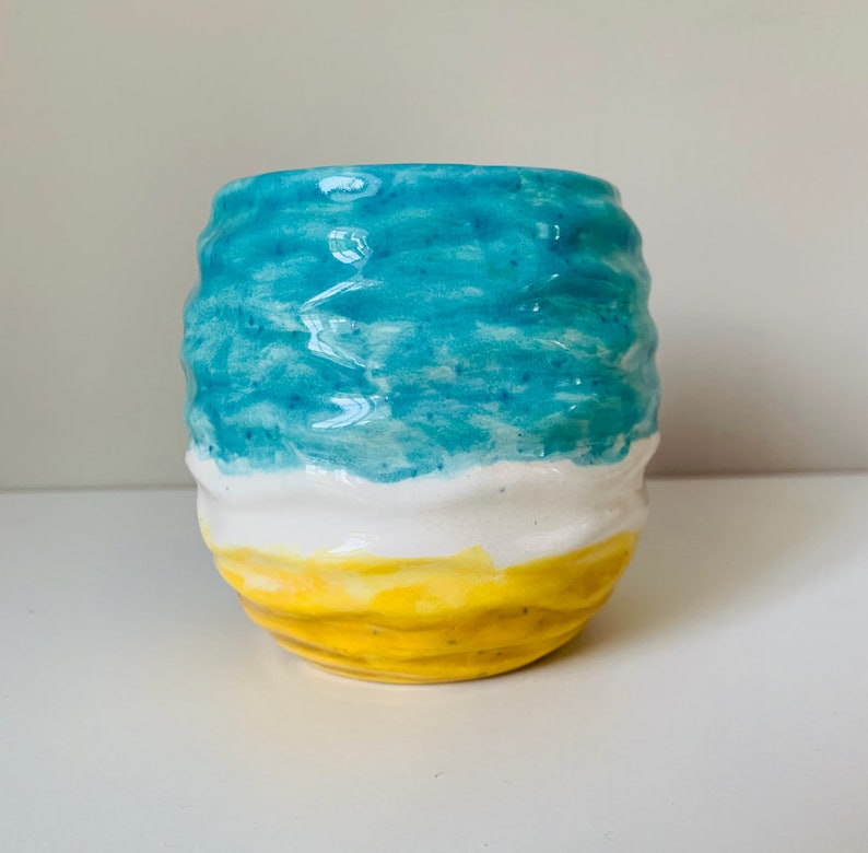 Puede incluir: Una taza de cer&aacute;mica con un esmalte azul, blanco y amarillo. La taza tiene una superficie ondulada y texturizada.